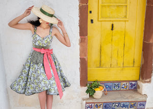 Frau steht vor einer Mauer neben einer gelben Türe mit einem Strohhut in grüngeblümten Kleid mit einem corallfarbenen Schleifenband . Auf der Stufe unter der Türe befindet sich ein kleiner Korb mit Zitronen und Orangen darin
