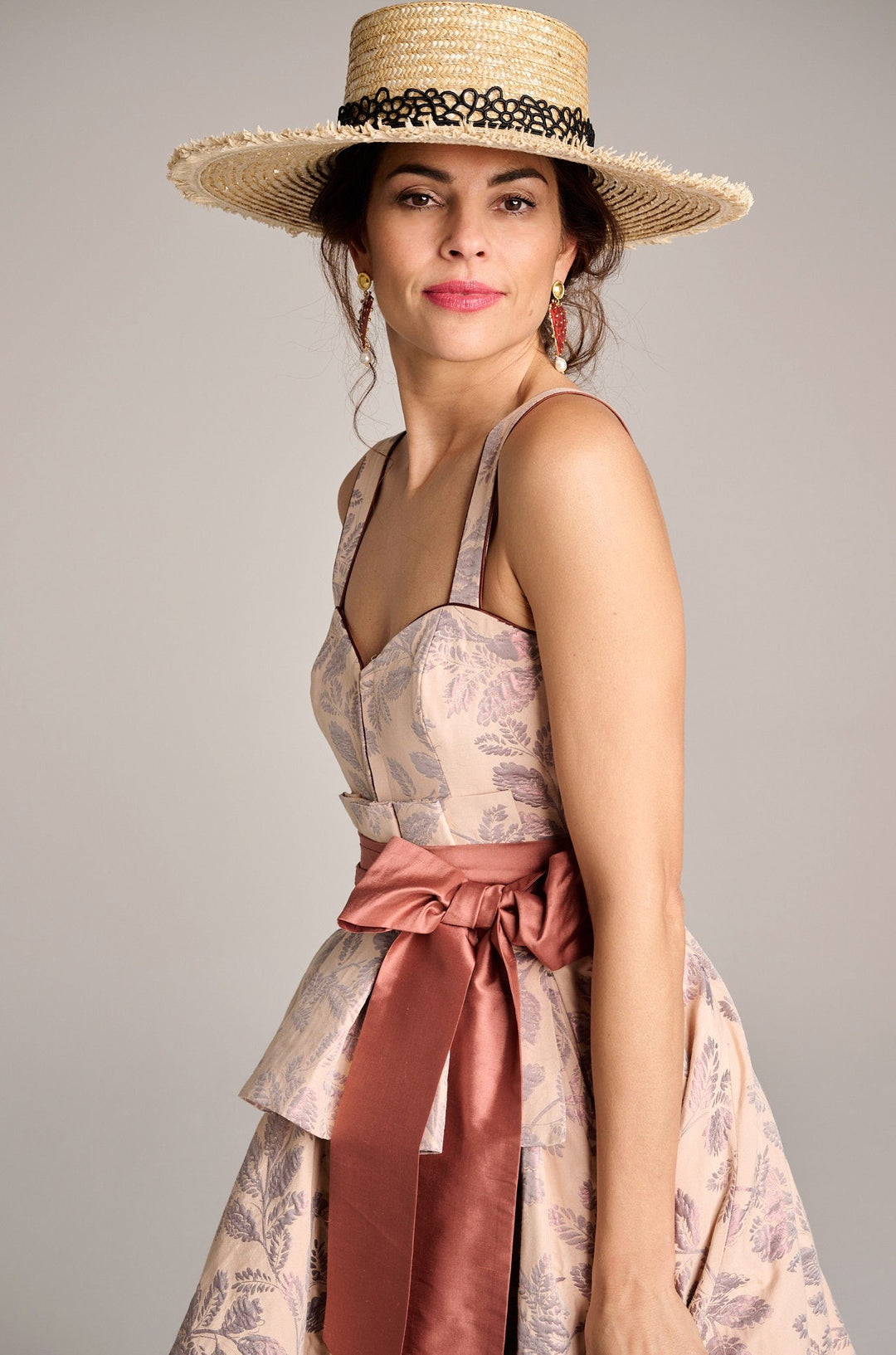 Eine schöne Frau im Designerdirndl in Rosa mit rougefarbenen Schleifenband trägt einen großen Strohhut während Sie selbstbewusst in die Kamera blickt