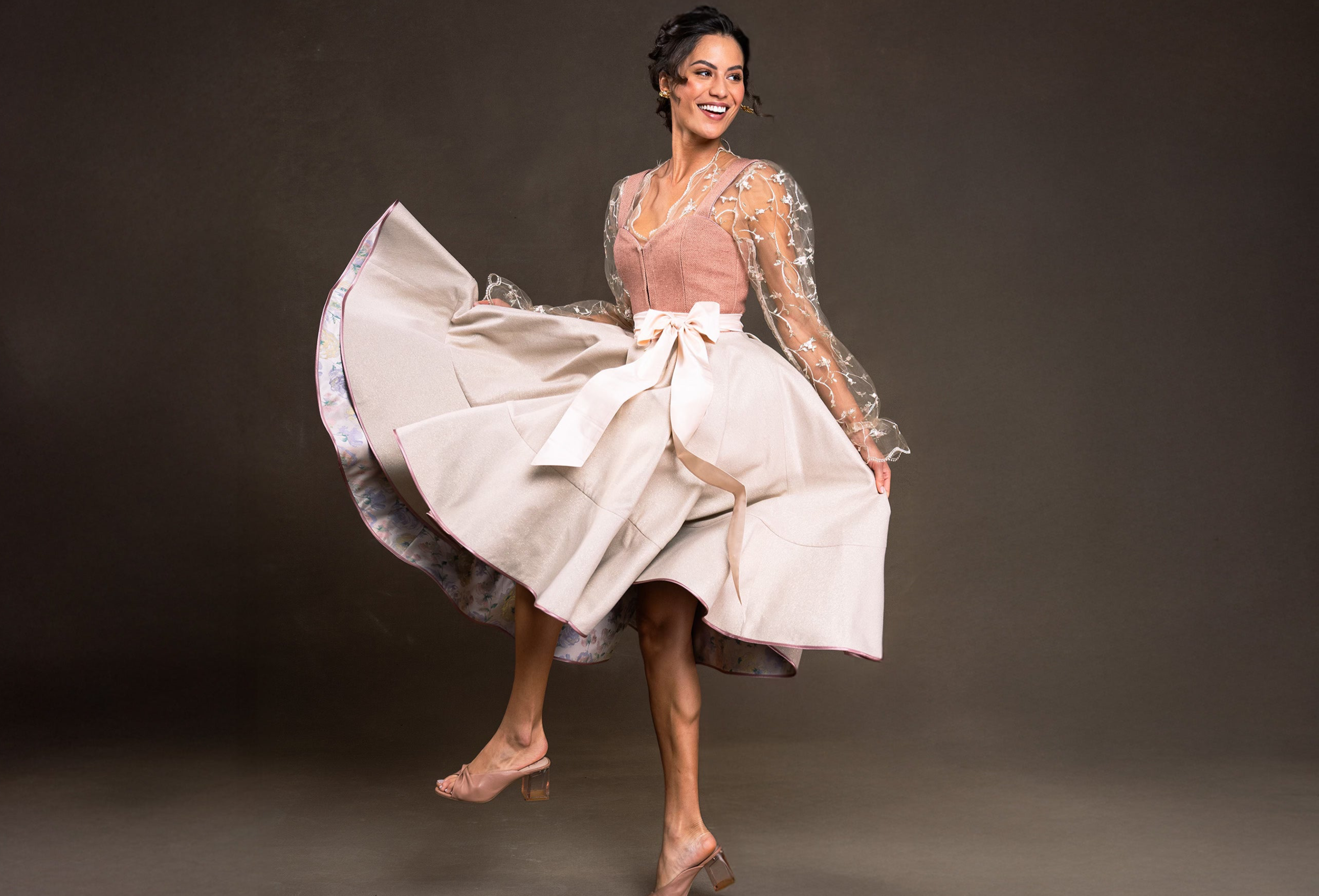 Dirndl springend und schwingend gezeigt von einer schönen Frau mit einer langarmigen transparenten Bluse in einem Altrosa tönen Kleid mit weissem Schleifenband